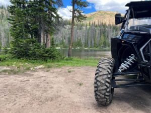 A close-up of the front left tire of a side-by-side UTV parked on a dirt trail with a lake and mountain behind it.