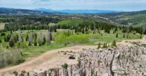 A dirt road curving along a rocky cliff with a small off-road vehicle, forests, and distant mountains beyond.