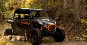 A front and side view of a grey and orange side-by-side UTV with driving on a dirt trail in the forest.