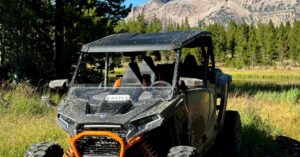 A frontal view of a dirty, side-by-side UTV with orange accents with a lake and mountain ridge behind it