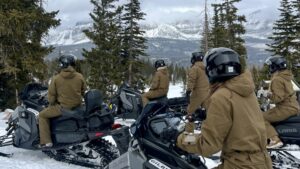 A rear view of a group of snowmobile riders wearing identical brown snowsuits parked and looking out at snowy mountains.