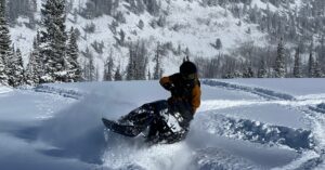An action shot of a person in a brown winter snowsuit and black helmet piloting a snowmobile through deep snow.