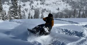 A person in a brown winter snow suit riding a snowmobile through deep snow on a ridge with a winter valley forest below.